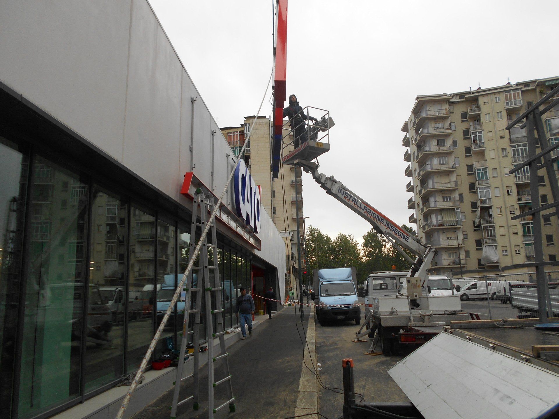 Operai che installano un cartello su un edificio con una piattaforma aerea. Camion e una scala sono visibili in un contesto urbano.