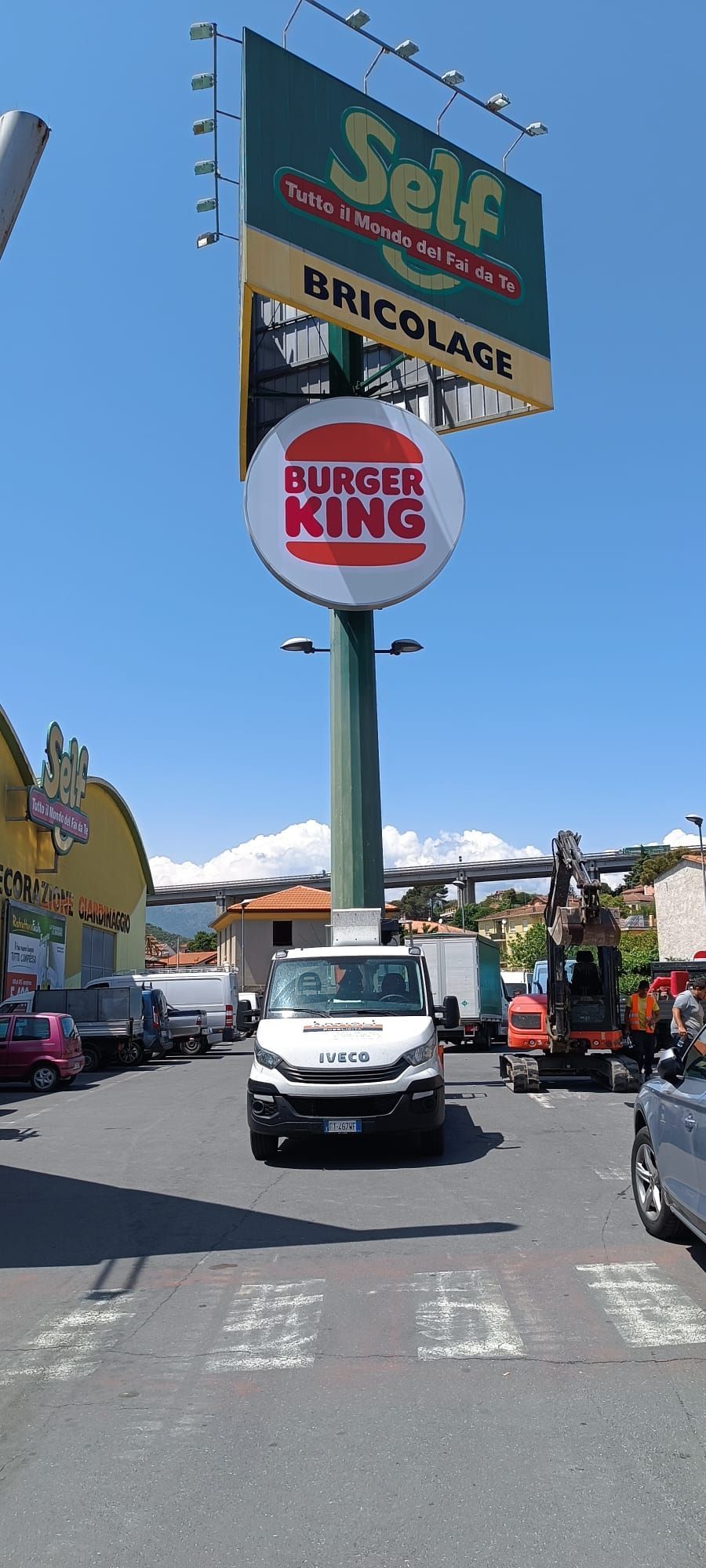 Un'insegna di Burger King su un palo alto di fronte a un negozio Self Bricolage con veicoli e cielo azzurro.