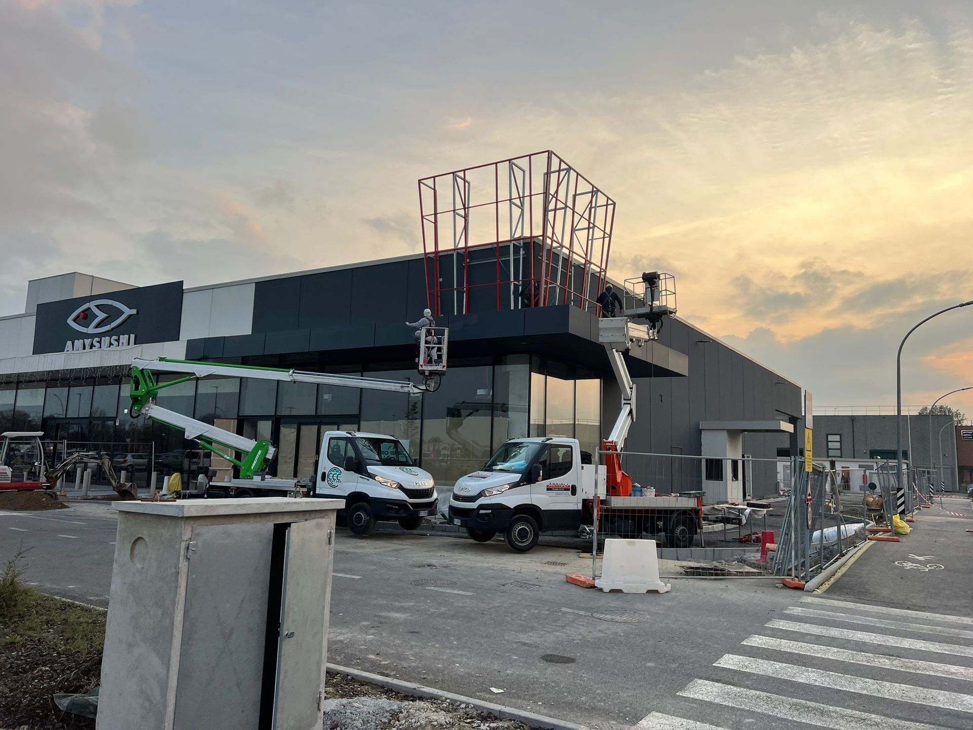 Cantiere con operai, camion e un cartello parzialmente costruito su un moderno edificio commerciale.