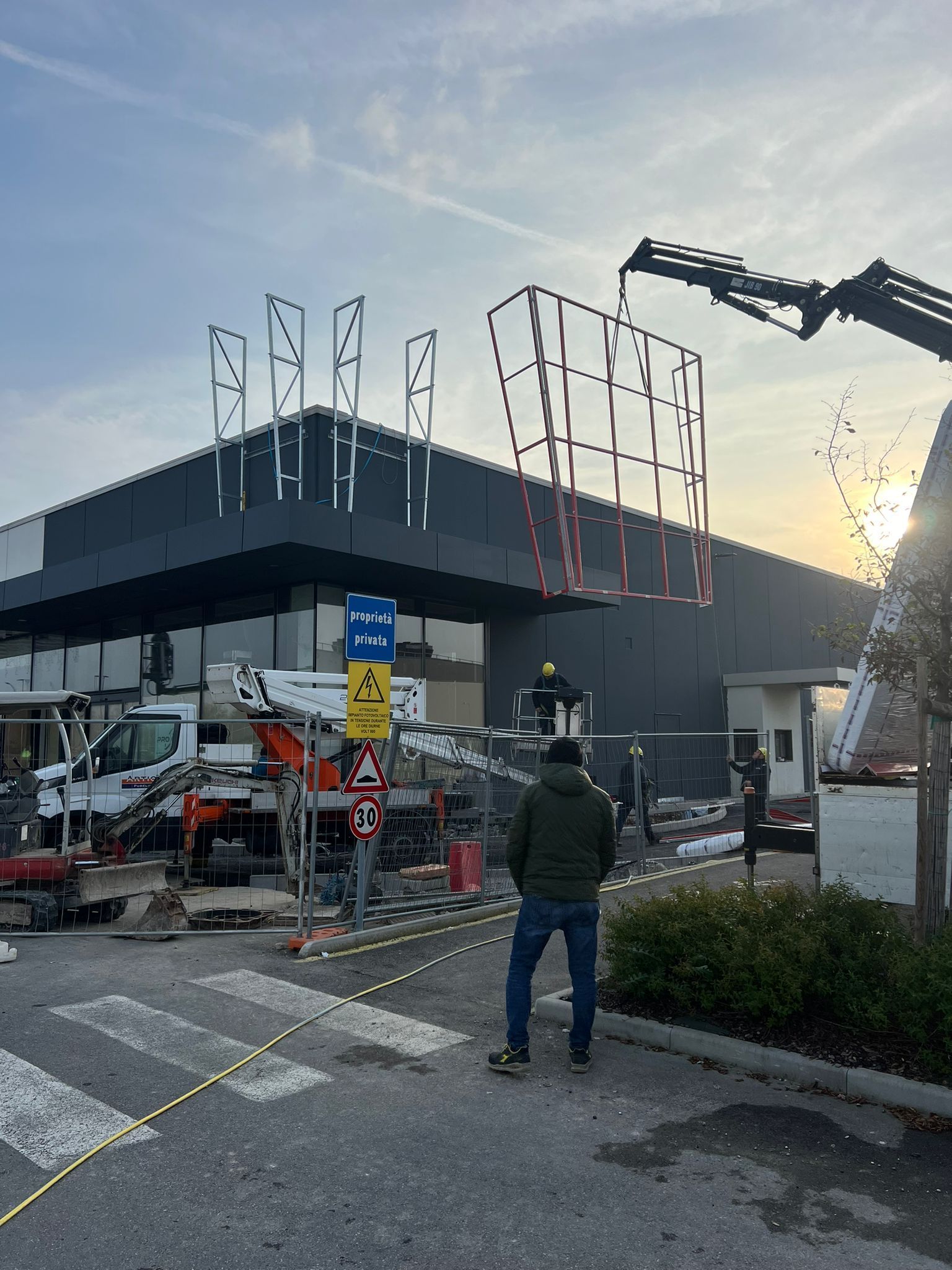 Cantiere edile con gru che solleva un cartello con struttura in metallo su un edificio buio; operai e un uomo osservano.