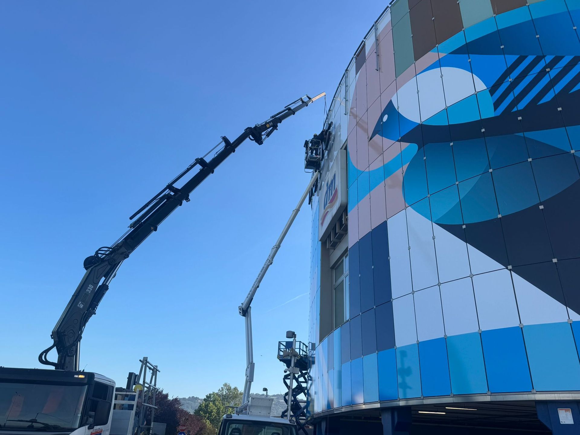 Due gru con operai che dipingono un grande murale colorato sulla facciata di un edificio sotto un cielo azzurro.