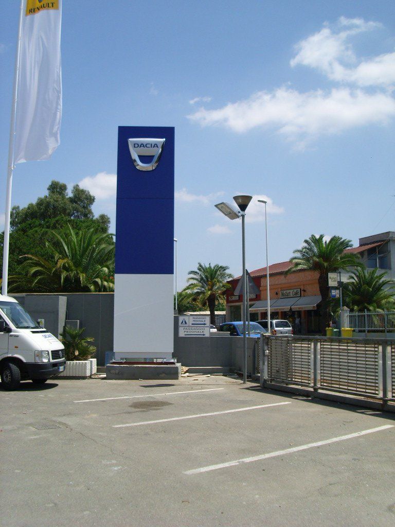 Insegna della concessionaria Dacia. Alto pilone blu e bianco con il logo Dacia. Auto e palme sullo sfondo.