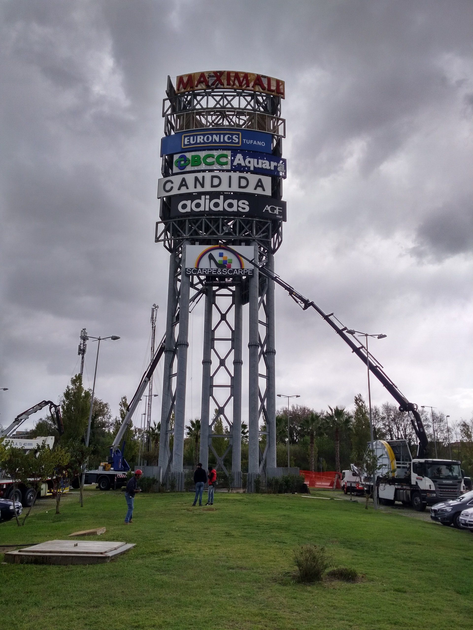 Una grande torre di insegne con diversi loghi aziendali, circondata da gru e operai in una giornata nuvolosa.