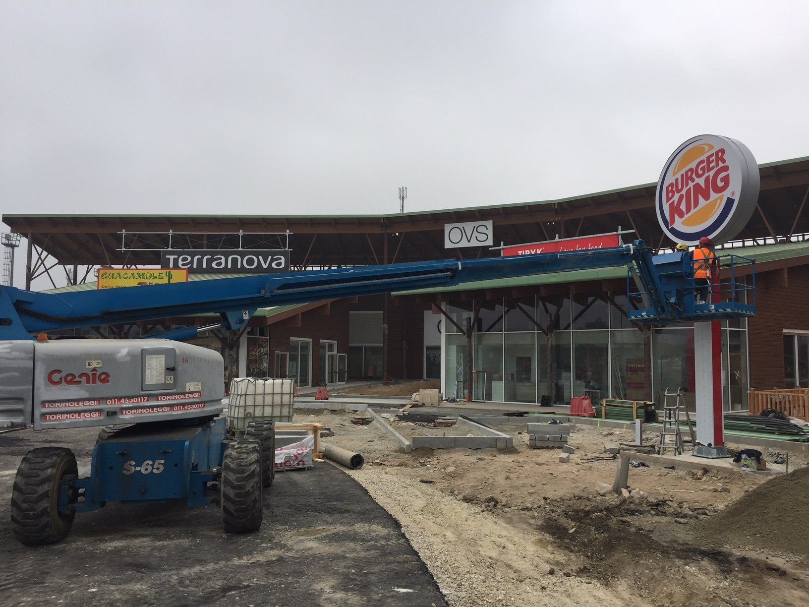 Cantiere con un'insegna di Burger King in fase di installazione. Carrello elevatore blu, cielo nuvoloso.