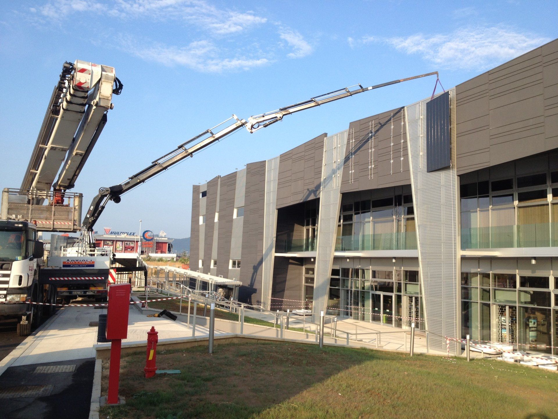 Un camion con un braccio gru esteso sta installando un cartello su un edificio moderno sotto un cielo azzurro.