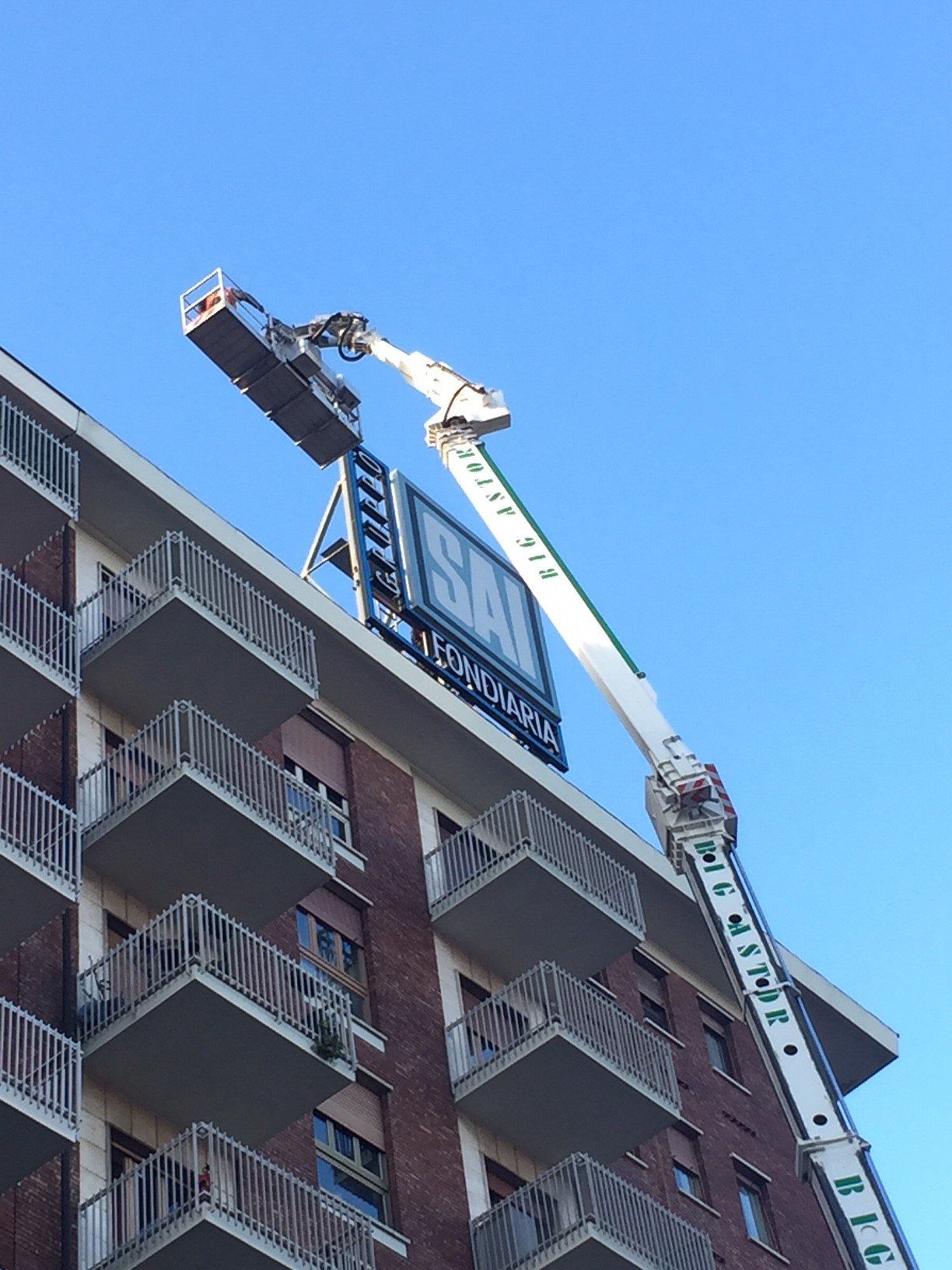 Una gru che solleva un'attrezzatura per installare un cartello su un edificio in mattoni. Cielo azzurro brillante.
