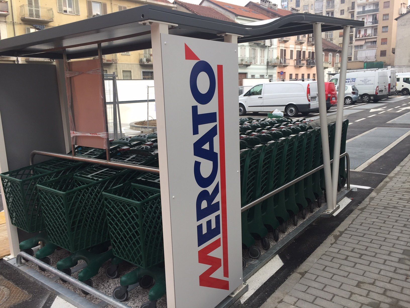 Recinto per carrelli della spesa del supermercato Mercato con carrelli verdi sotto una tettoia, parcheggiati su un marciapiede, cartello sul lato.