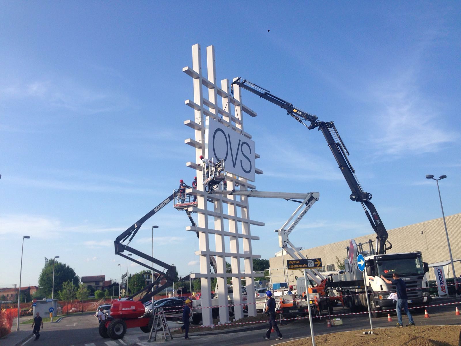 Operai edili installano l'insegna di un negozio OVS, con gru sullo sfondo del cielo azzurro.