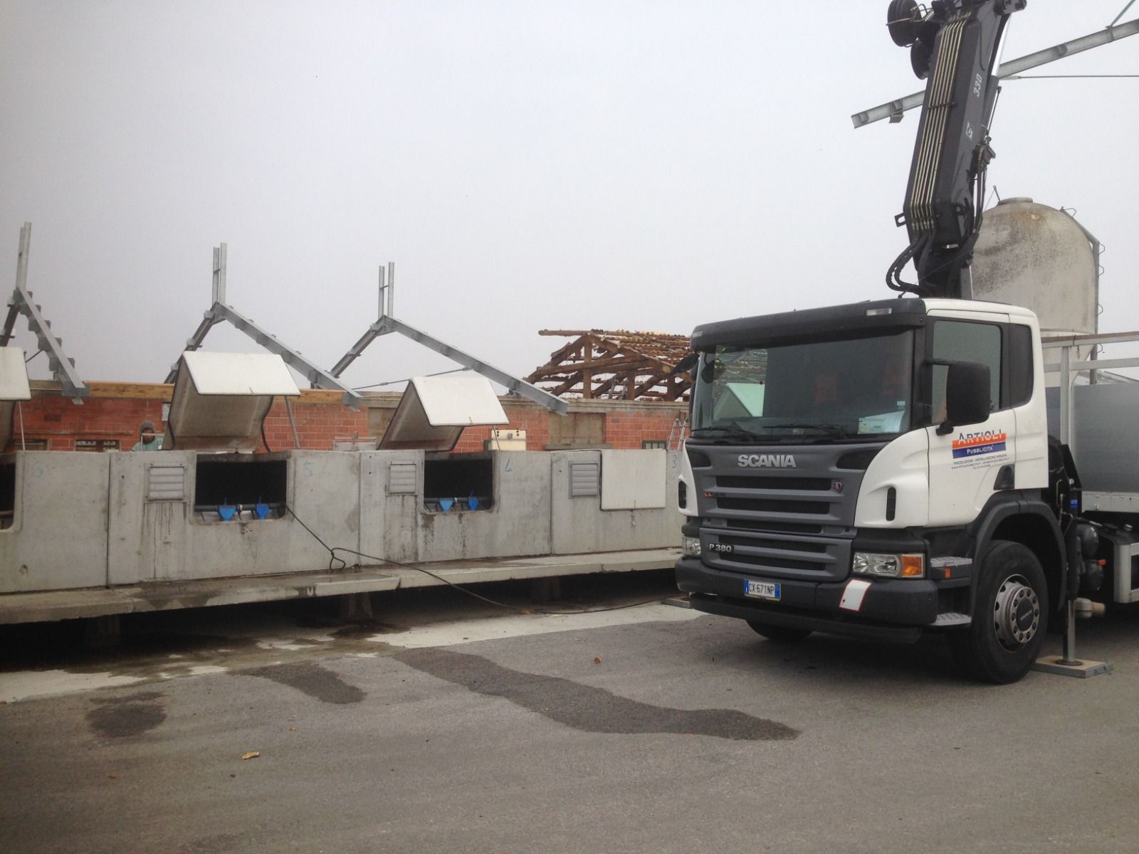 Un camion Scania con una gru accanto a una struttura in cemento in costruzione in una giornata grigia.