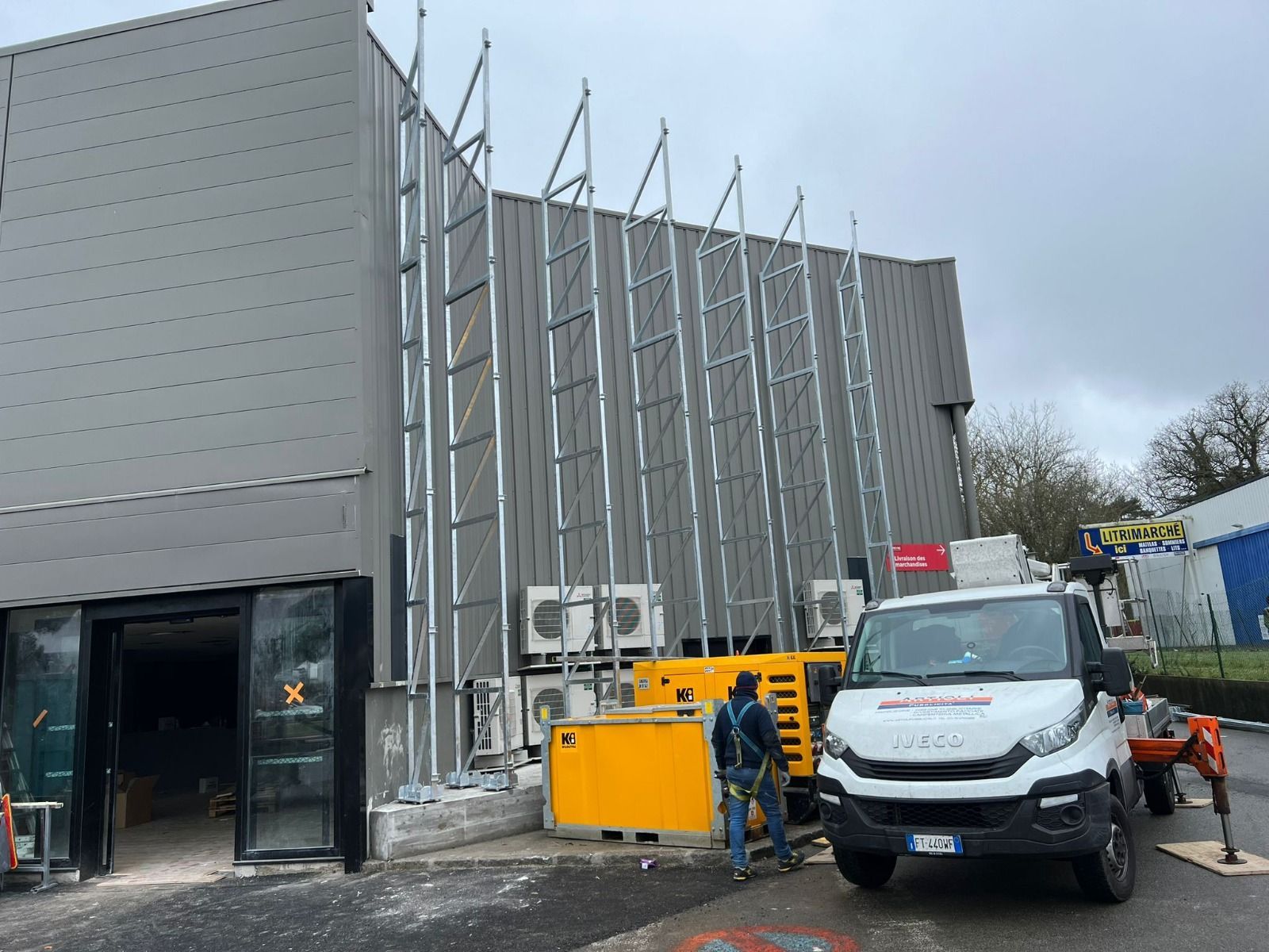 Cantiere con impalcature sul lato di un edificio grigio, un generatore e un furgone bianco.