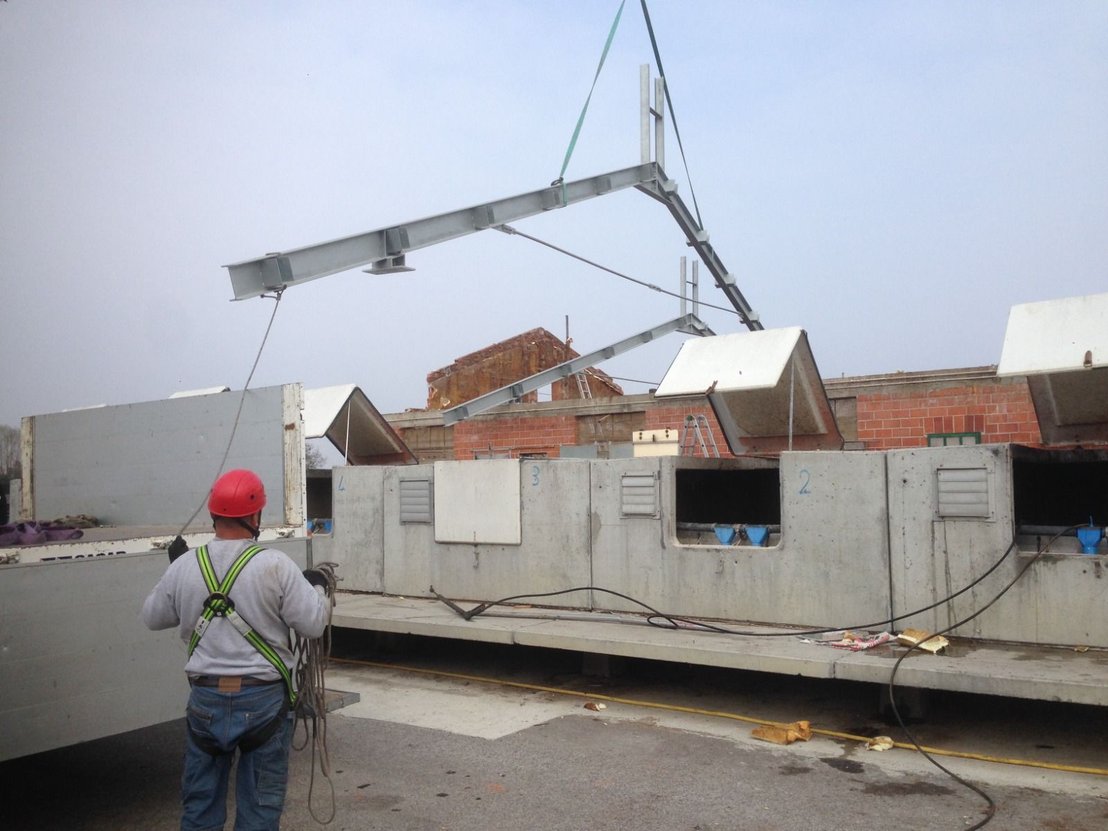 Operaio edile che guida un pannello di cemento con una gru, sullo sfondo un edificio in mattoni. Cielo nuvoloso.