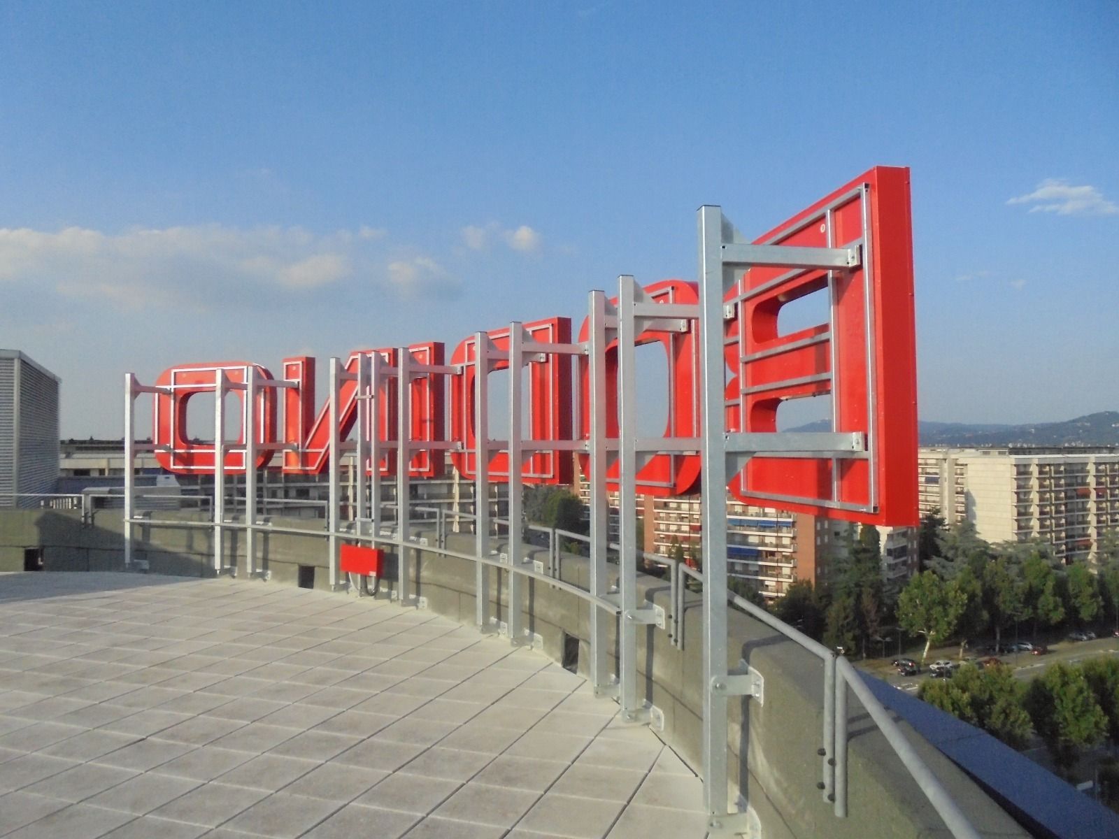 Un cartello rosso e argento su un tetto che domina un paesaggio urbano. Sembra che il cartello scriva una parola.