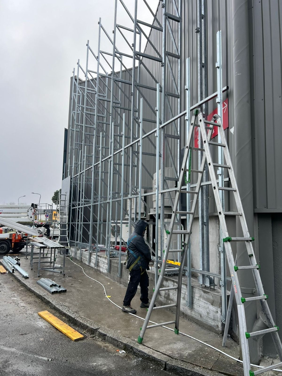 Operaio edile vicino a un'impalcatura metallica sul lato dell'edificio; scala, esterno grigio, cielo coperto.