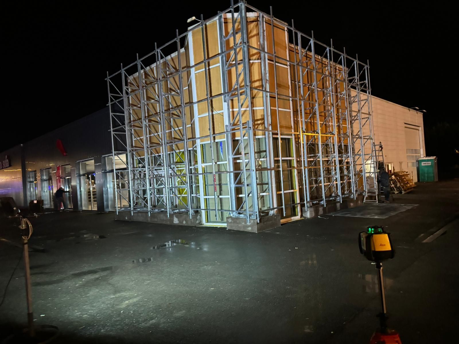 Esterno di un edificio in costruzione di notte, con struttura metallica, livella laser gialla e personale addetto alla costruzione.