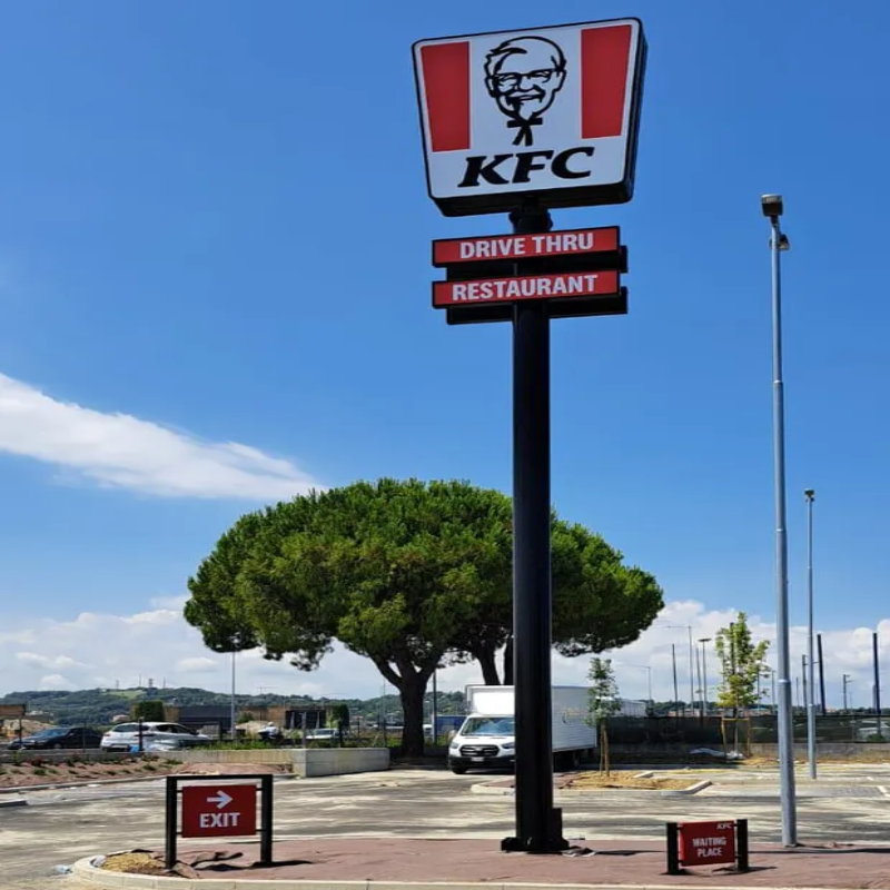 Cartello KFC con opzioni di drive-thru e ristorante, palo nero, cielo azzurro.