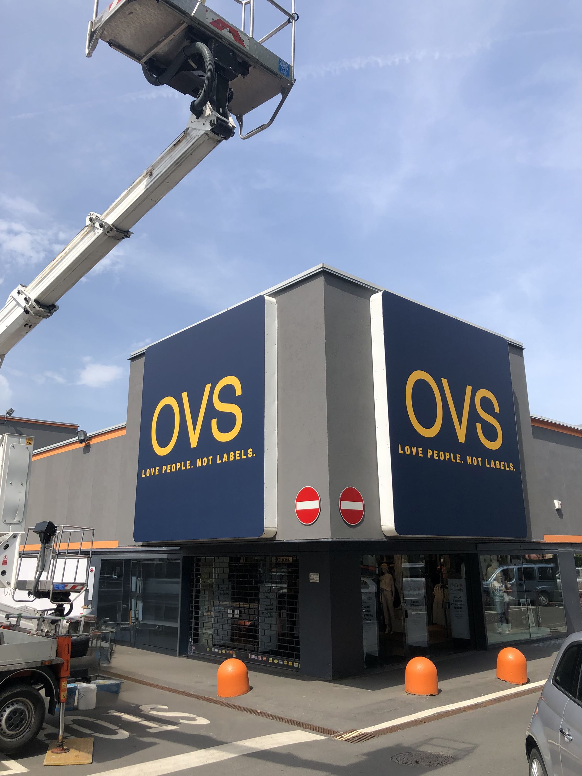 Esterno del negozio OVS con la segnaletica in fase di realizzazione da un ascensore, sotto un cielo azzurro.