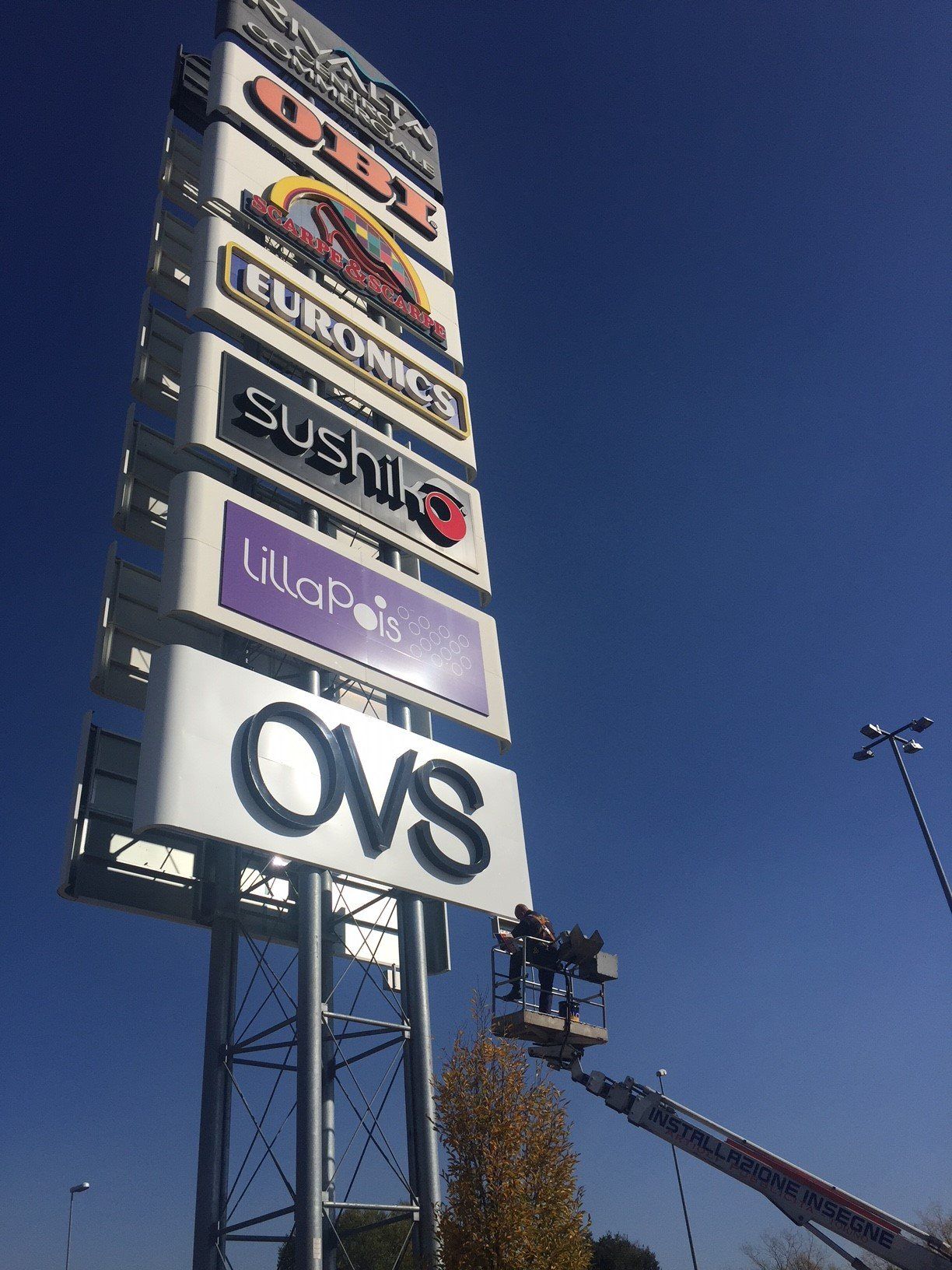 Grande cartello di un negozio con loghi, su cui lavorano persone in ascensore; cielo azzurro e limpido.