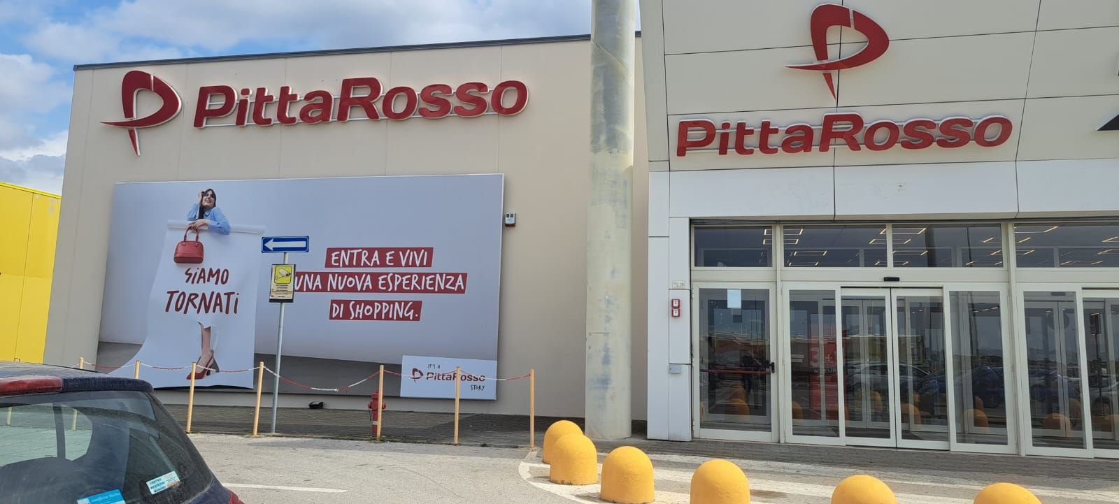 Esterno del negozio PittaRosso con grande logo rosso. Pubblicità sul muro. Porte automatiche visibili.