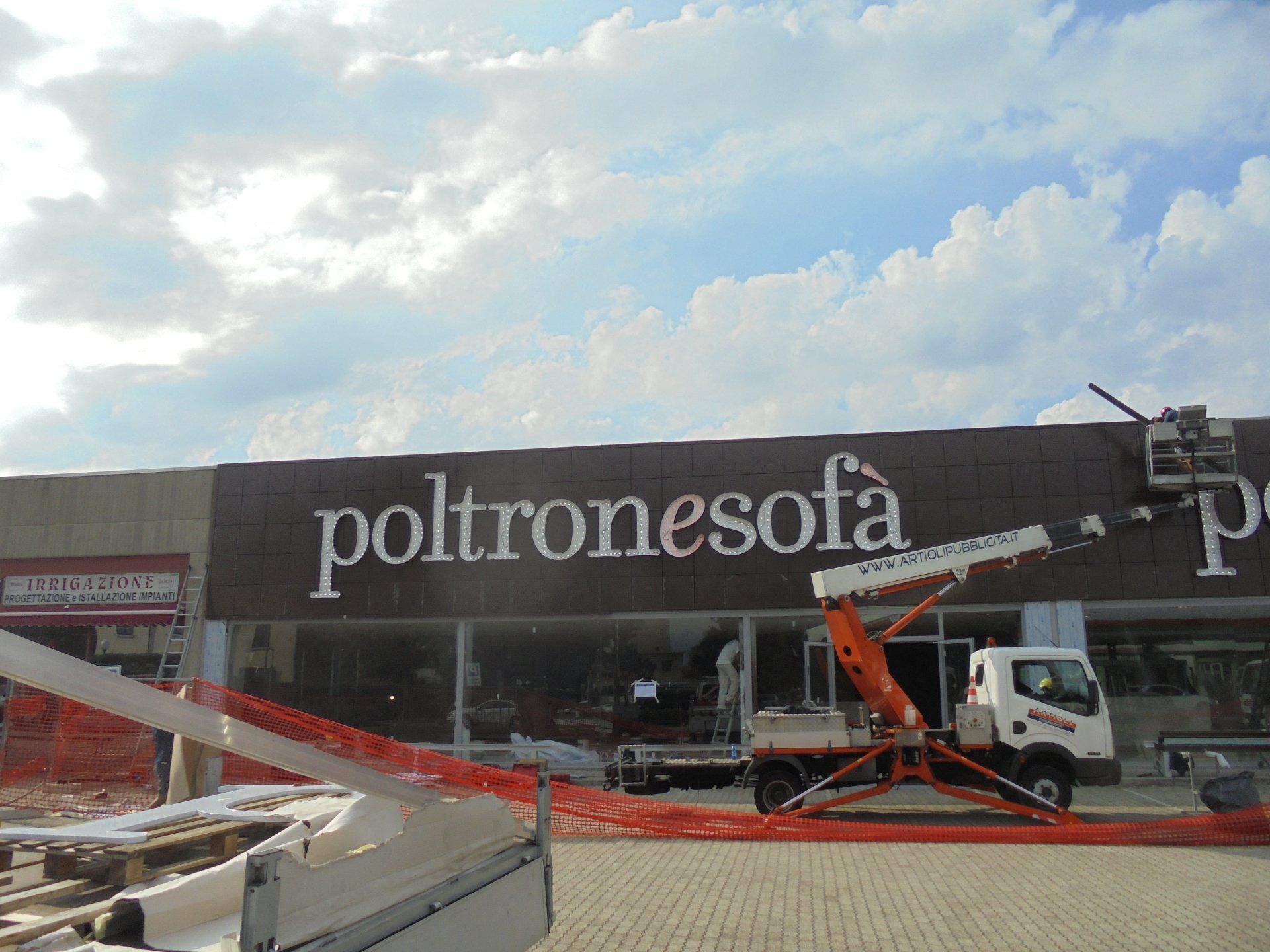Installazione della segnaletica presso un negozio Poltronesofa, con un sollevatore montato su camion e cielo nuvoloso.