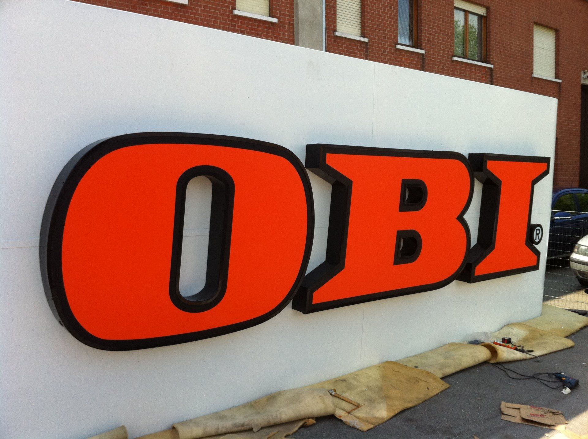 Insegna OBI: lettere arancioni con rifiniture nere su una superficie bianca, probabilmente per un negozio.