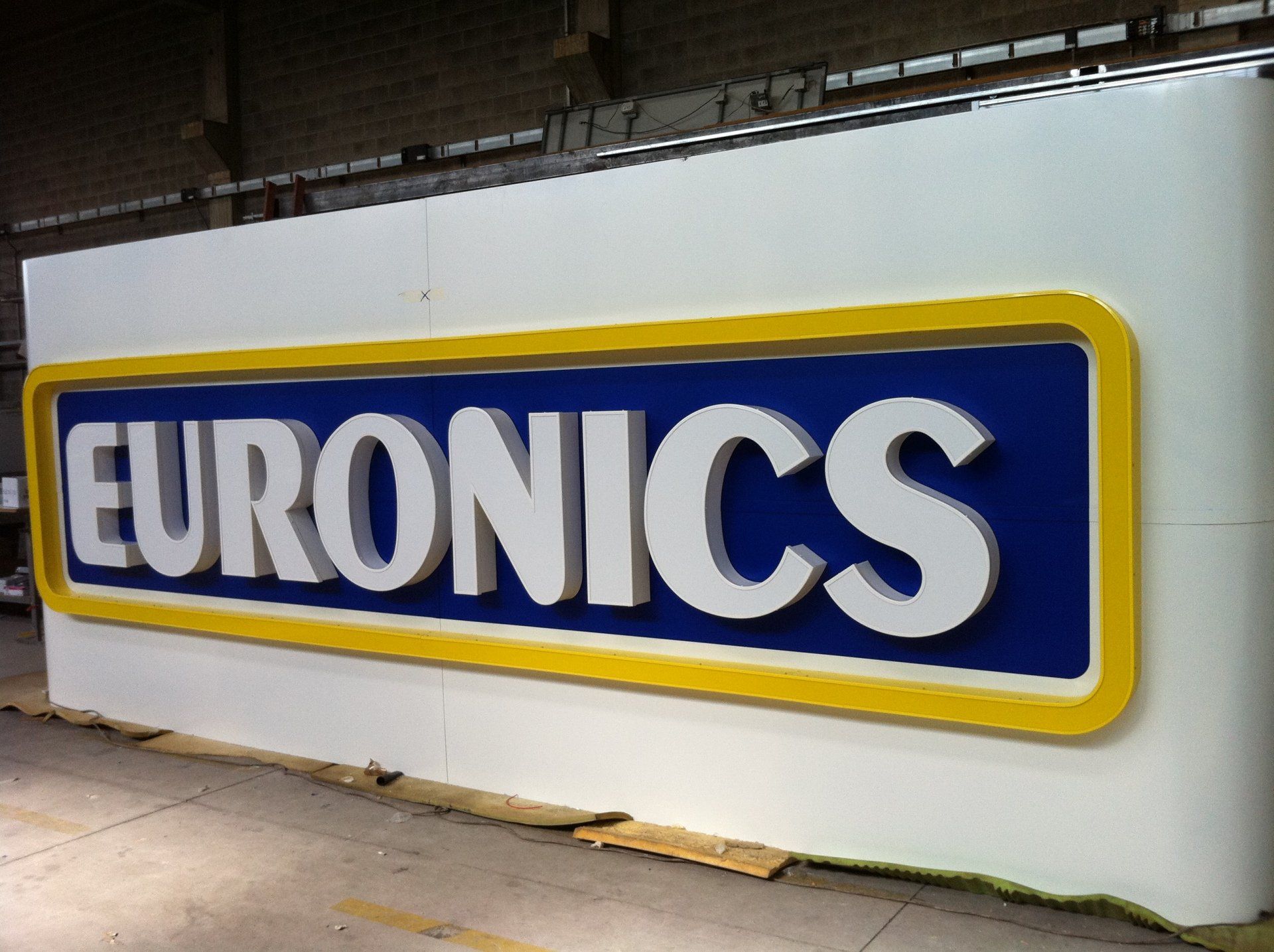 Grande cartello Euronics con lettere bianche su sfondo blu e bordo giallo, montato su una parete bianca.
