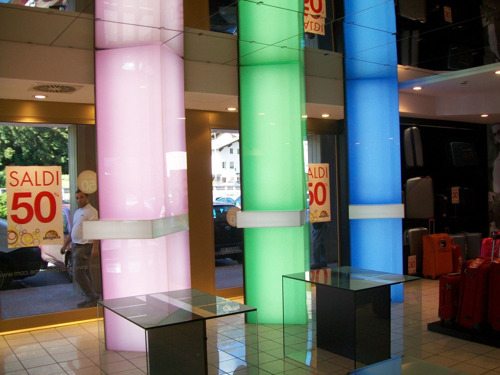Tre colonne colorate (rosa, verde, blu) ben illuminate in un negozio, con espositori in vetro e un cartello con la scritta 