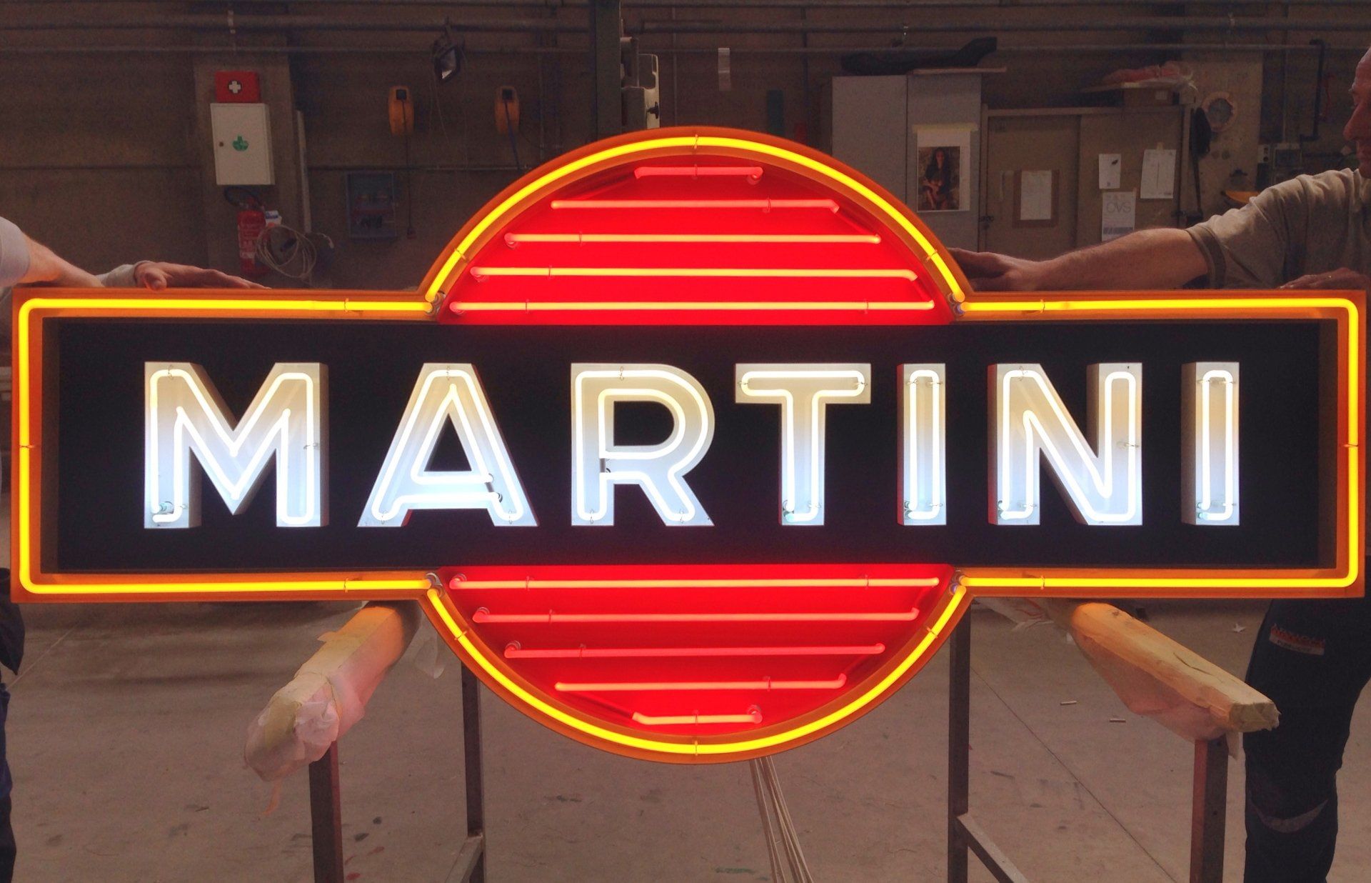 Insegna al neon Martini con contorno giallo e testo bianco. Tenuta da due persone in un laboratorio.