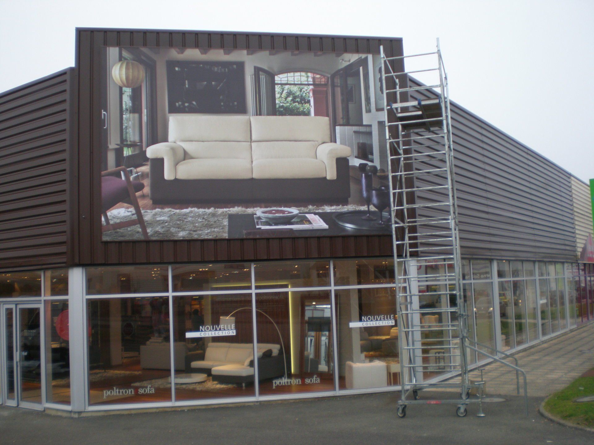 Esterno di un negozio di mobili in vetro e marrone con un grande divano pubblicitario e un'impalcatura.