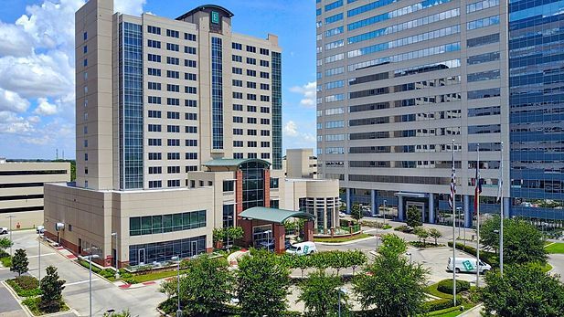 A large building with a lot of windows is surrounded by trees and a parking garage.