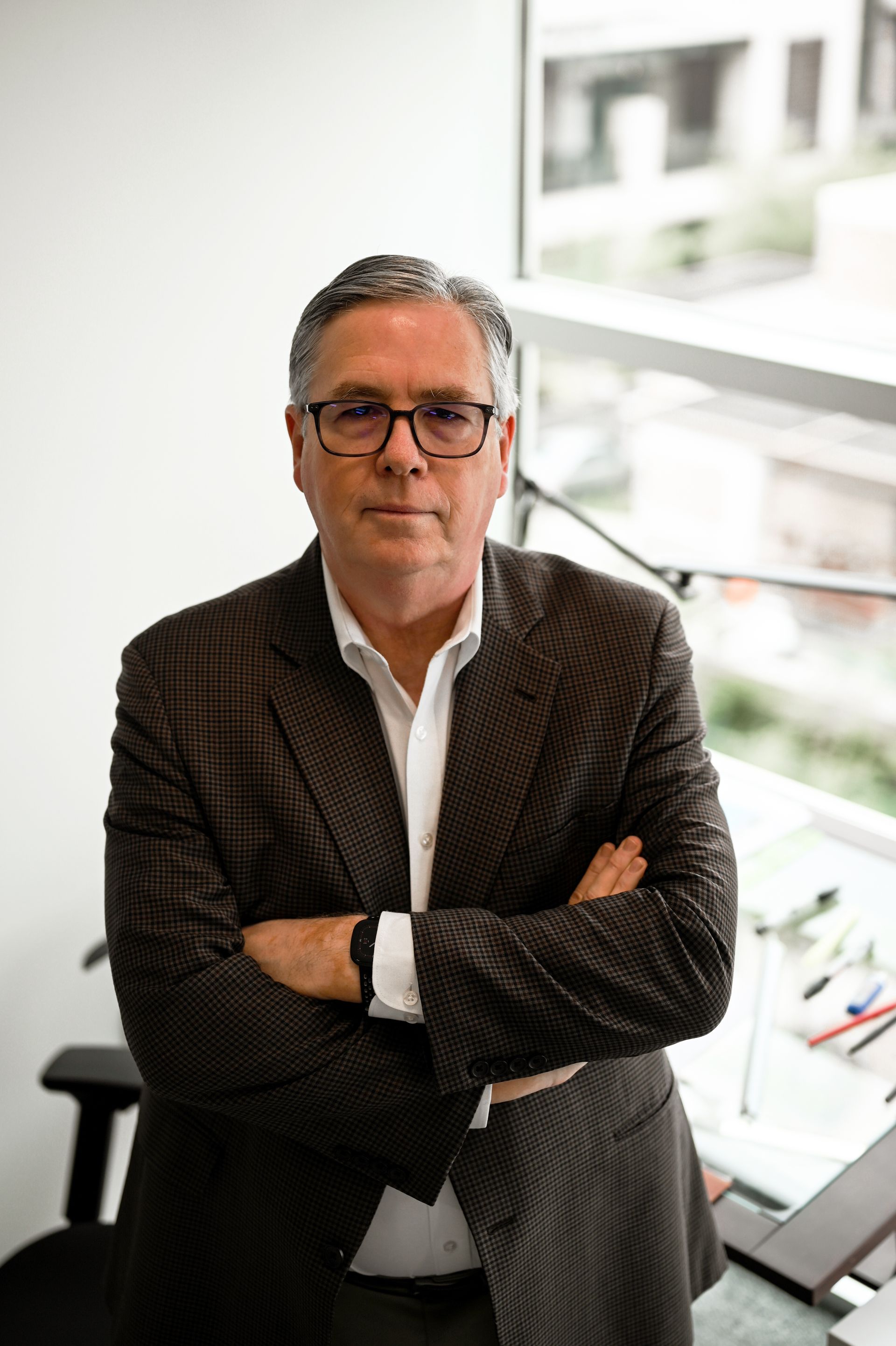 A man in a suit and glasses is standing in front of a window with his arms crossed.