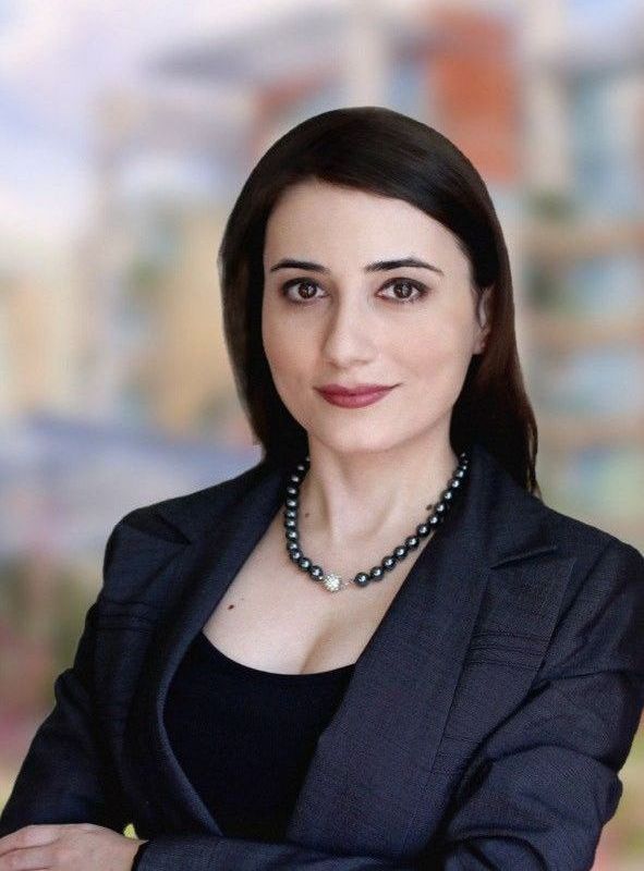 A woman in a suit and necklace is standing with her arms crossed.