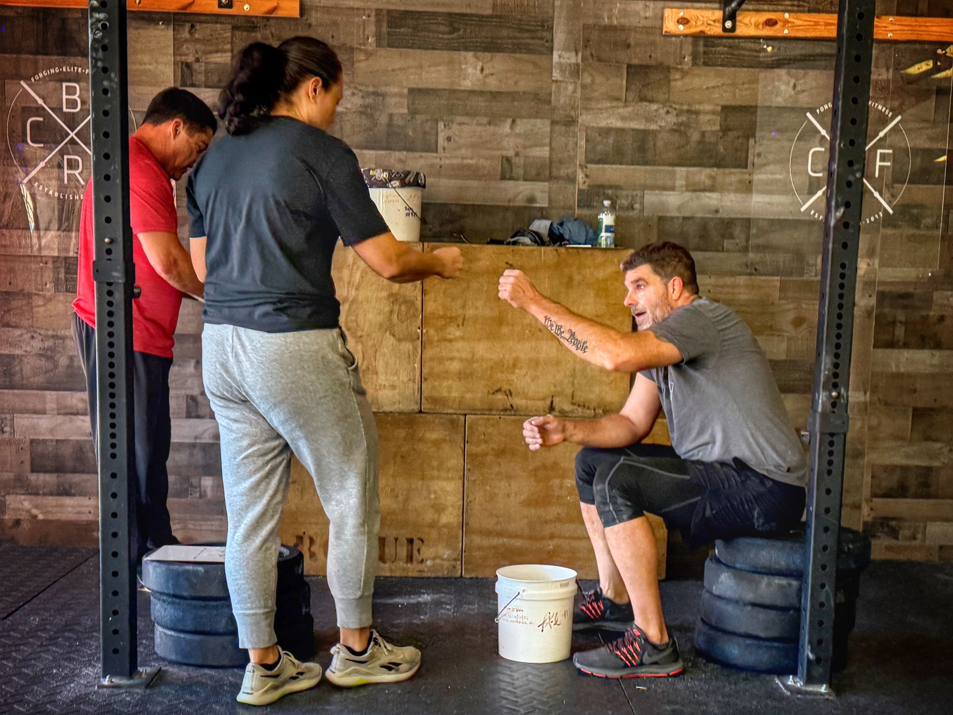 People in a gym, one seated, gesturing. Others are standing by equipment.