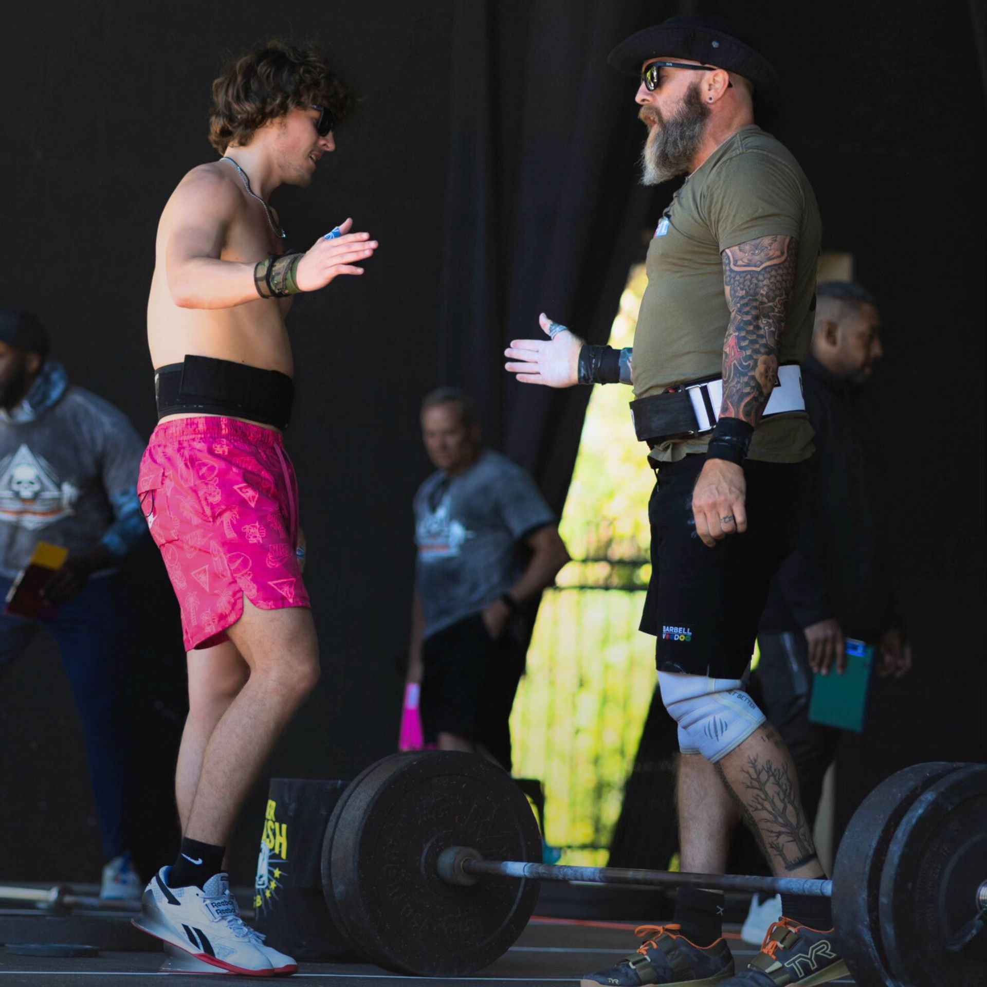 Two men in athletic gear high-fiving near a barbell. Outdoor setting, sunny.