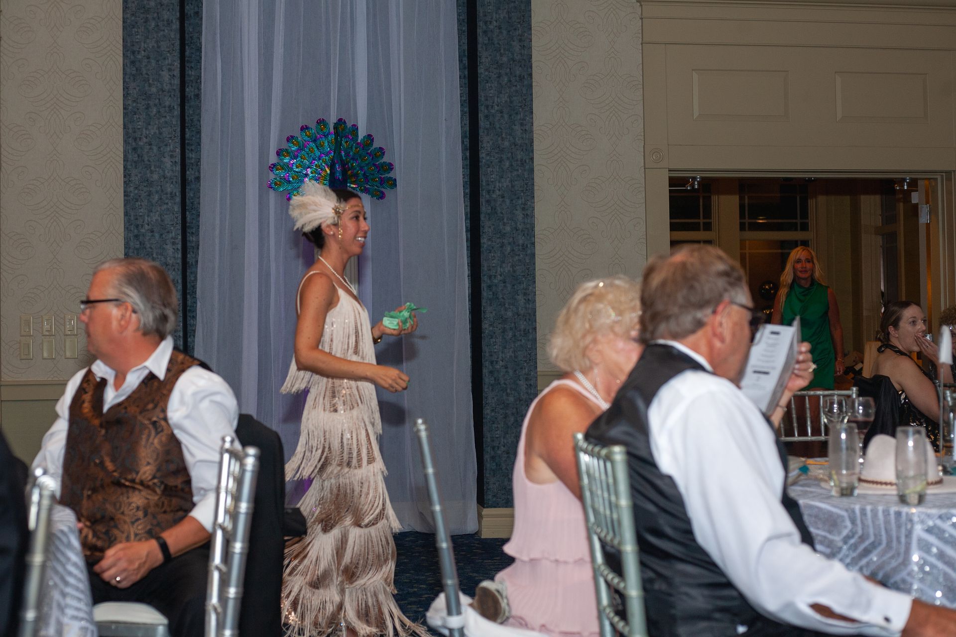 A woman in a dress and peacock hat is standing in front of a group of people sitting at tables.