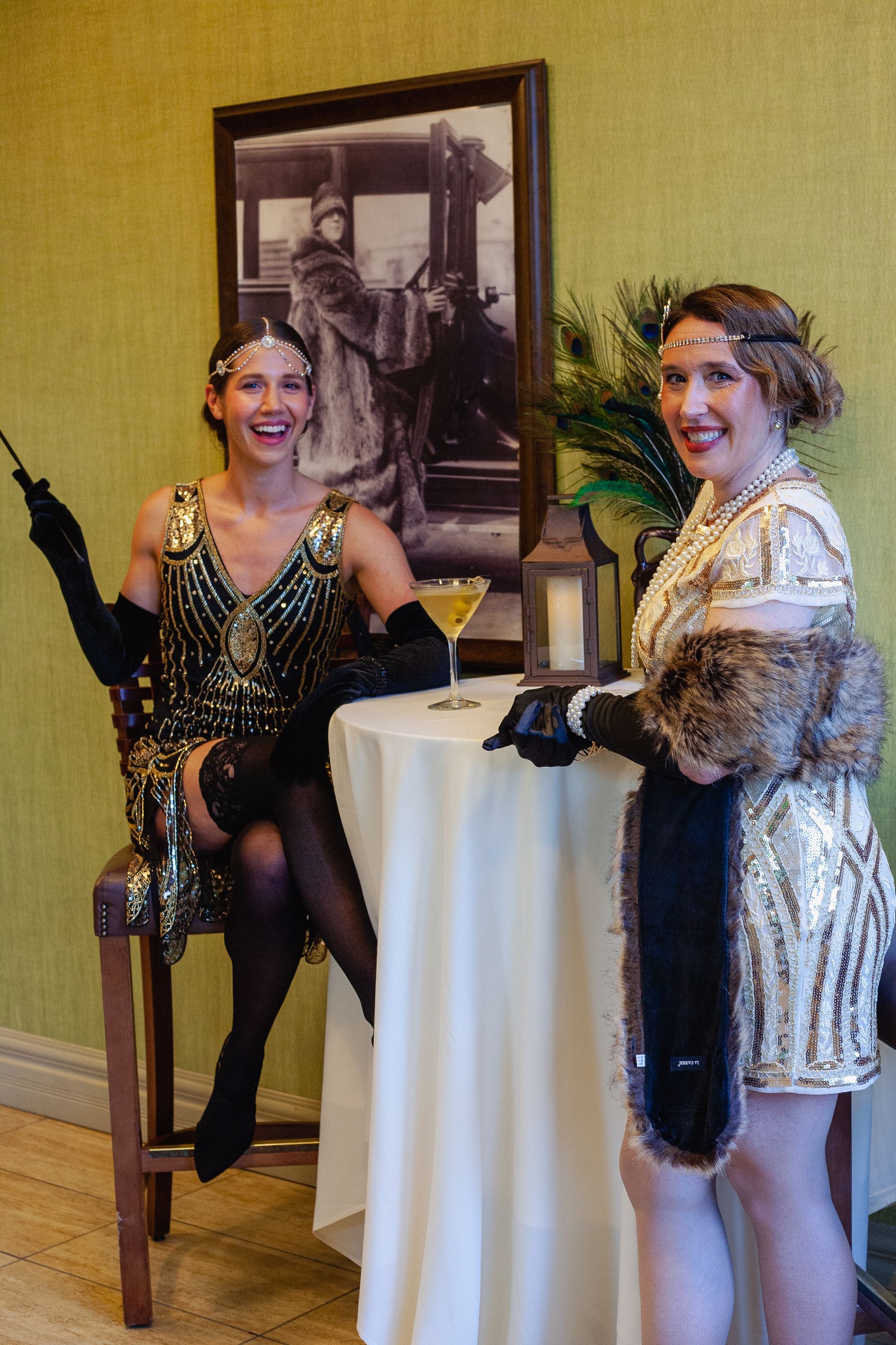 Two women in flapper dresses are sitting at a table with a martini.