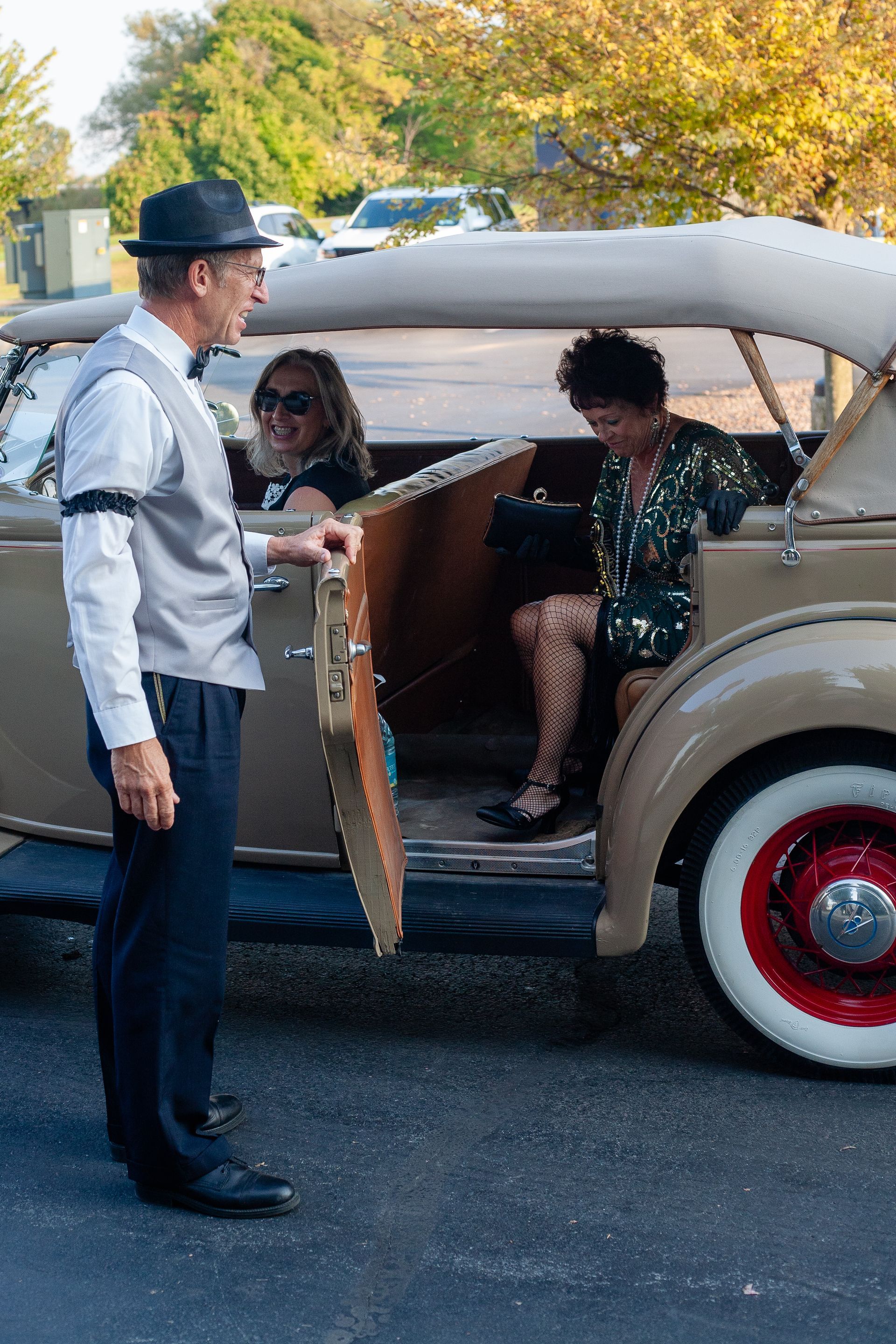 A man in a hat is standing next to a car with two women in it.