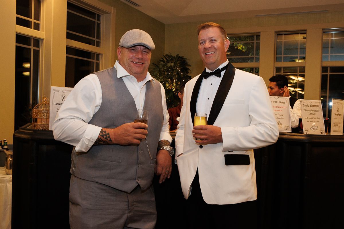 Two men in tuxedos are standing next to each other holding drinks.
