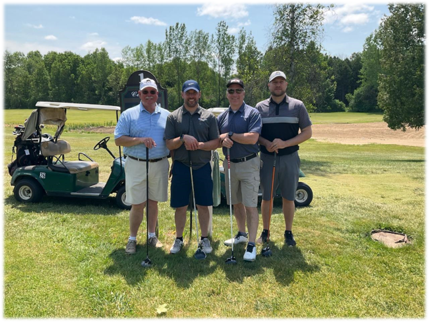 Four men are standing on a golf course with golf clubs in front of a golf cart.