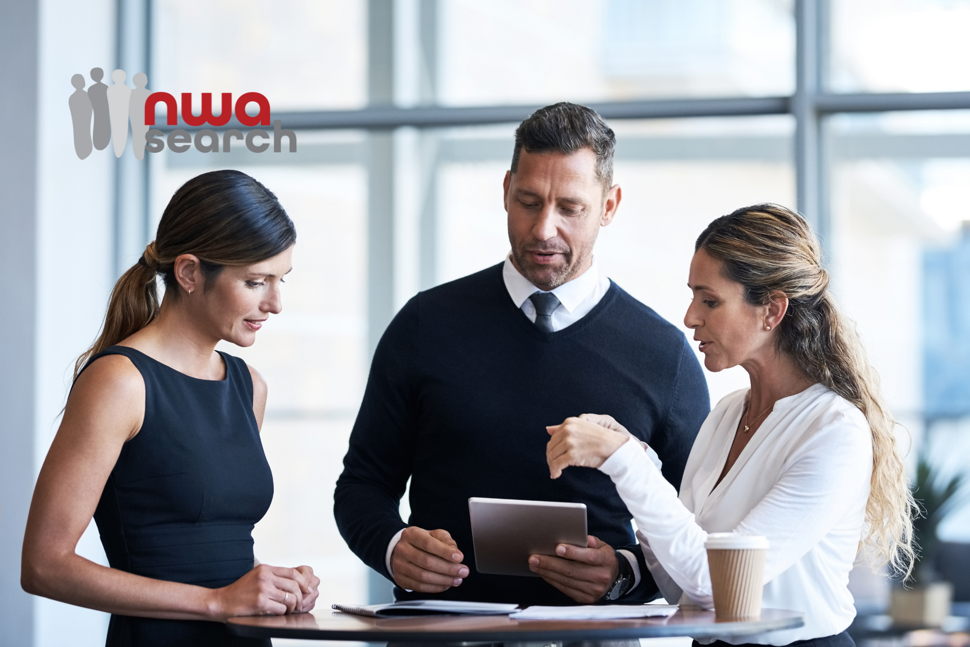 A group of people are standing around a table looking at a tablet discussing hiring trends 2025