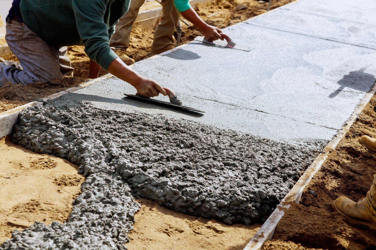 Workers smoothing wet concrete for a sidewalk, framed by wooden borders, set in sandy ground.
