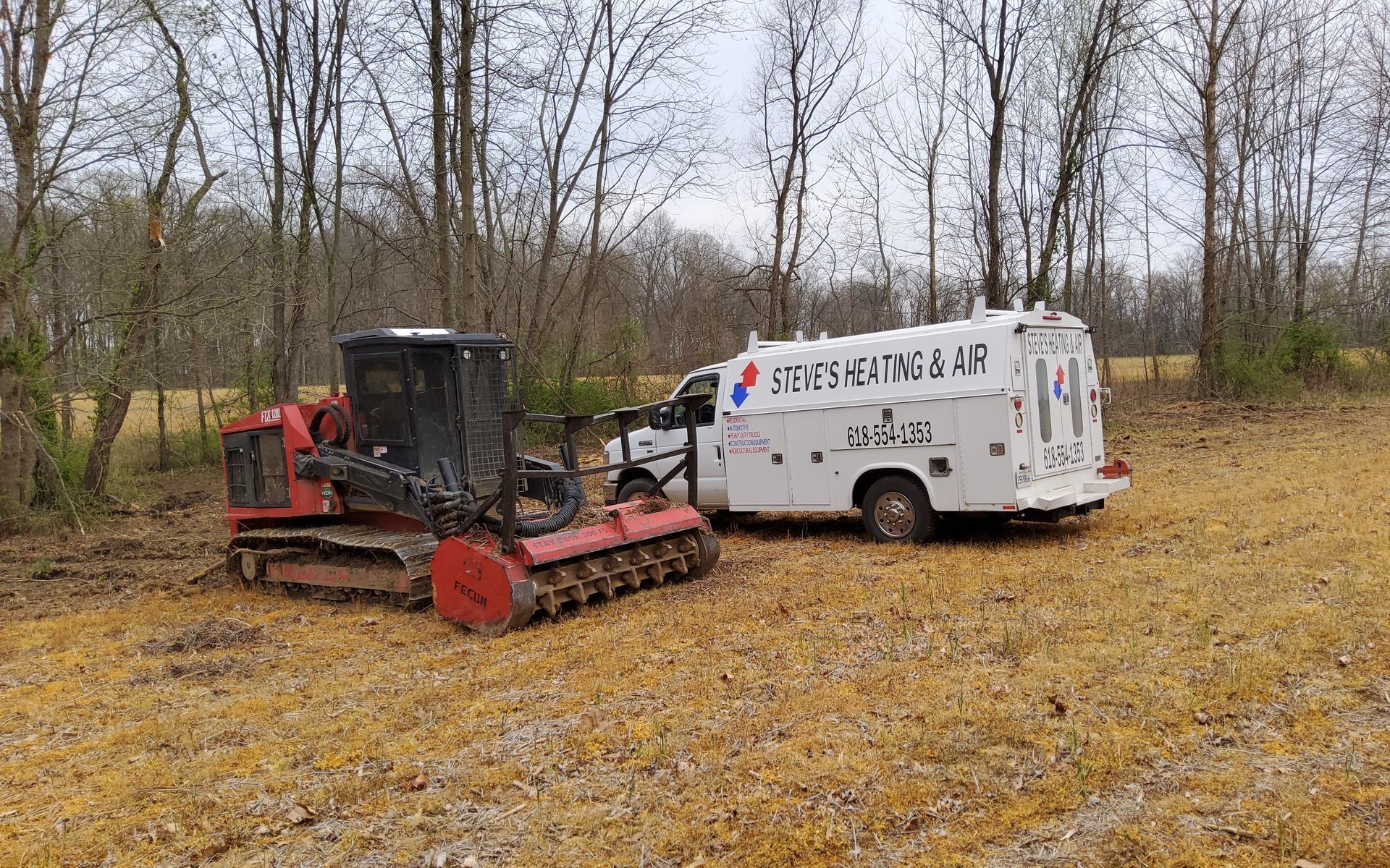 A track-mounted forestry mulcher next to a service truck, both on a dirt path in a wooded area.