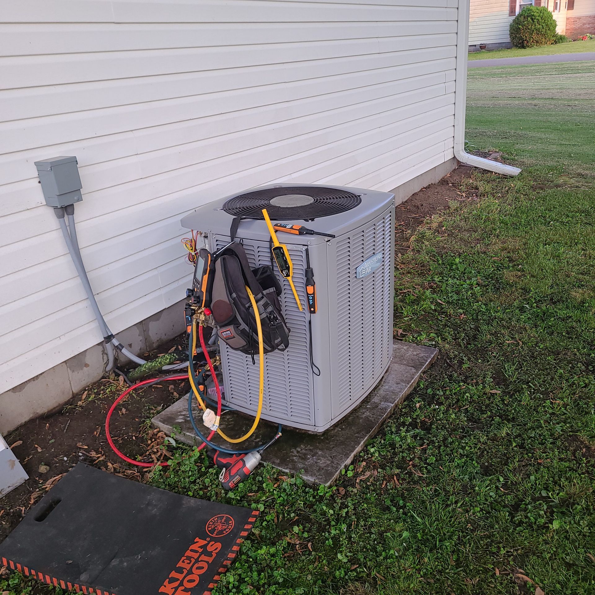 HVAC unit being serviced outdoors, with tools connected. Grey unit, white siding, and green grass.