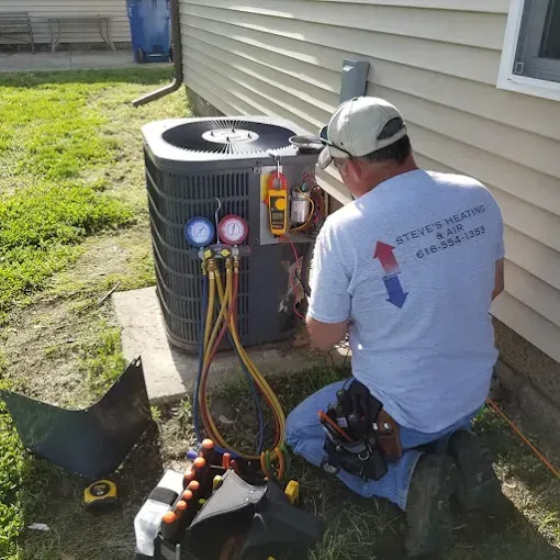 HVAC technician kneels beside an air conditioning unit, using gauges and tools. Outside near a house.