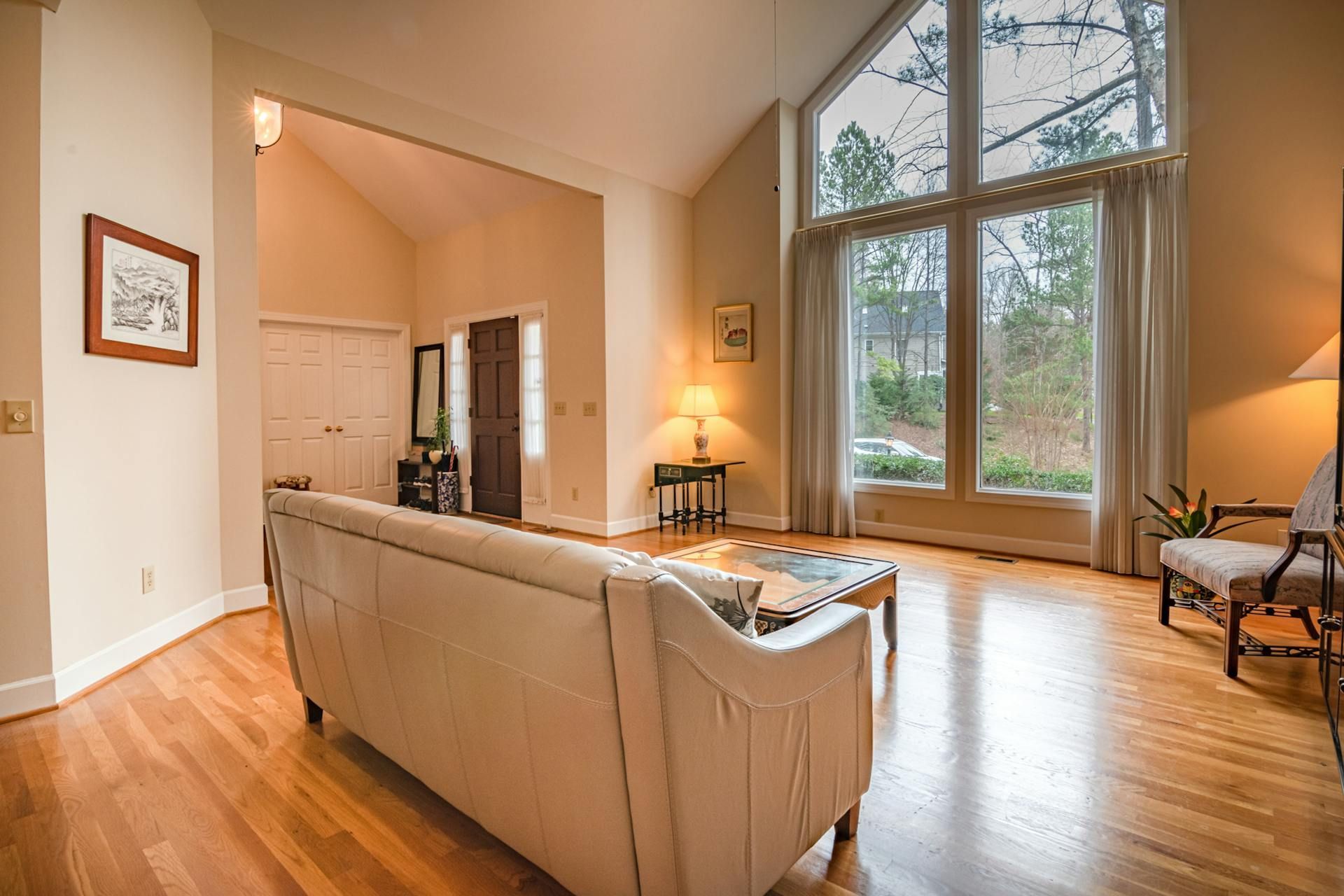 A sunlit living room with hardwood floors, a beige sofa, and a large window looking out onto a wooded area.