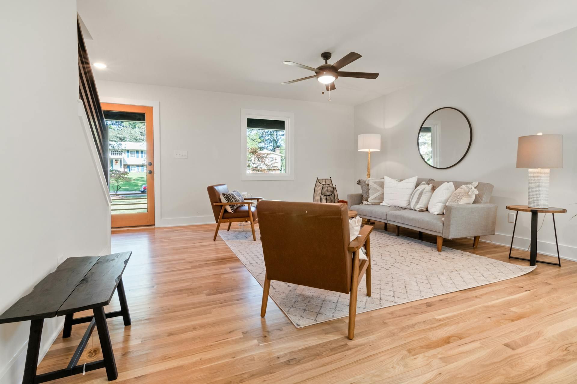 A bright living room with light wood floors, a gray sofa, brown leather accent chairs, a round mirror, and a wood door.