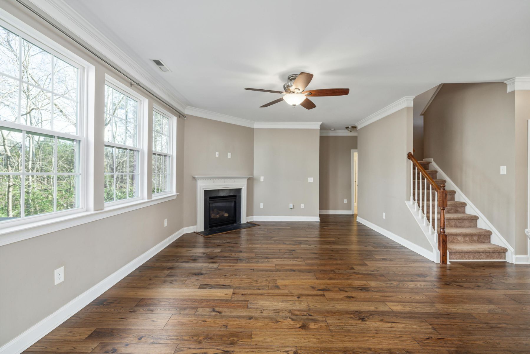 A spacious living room featuring hardwood floors, a fireplace, large windows, and a staircase leading to an upper level.