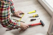 Person measuring a floorboard with a tape measure beside a hammer and tools