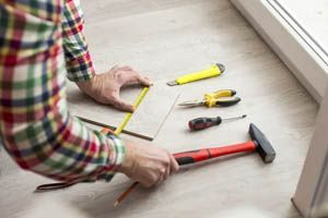Person measuring a floorboard with a tape measure beside a hammer and tools