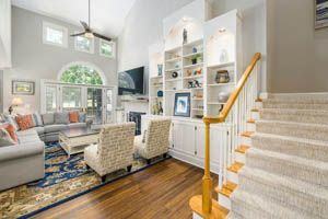 Bright living room with gray sofa, woven chairs, blue rug, built-in shelves, and staircase with wood handrail