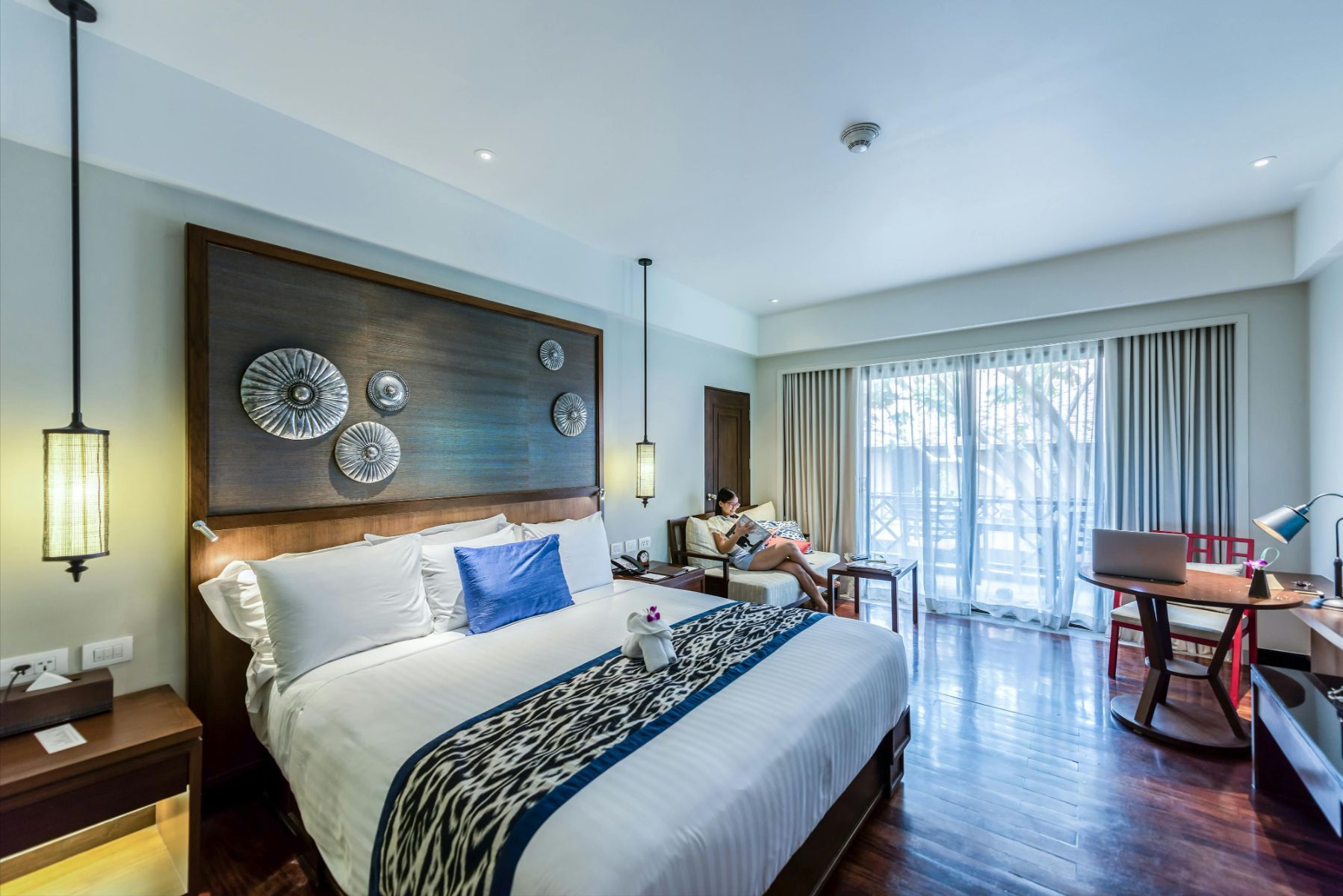 Modern hotel bedroom with king bed, blue accents, desk, and balcony doors overlooking trees
